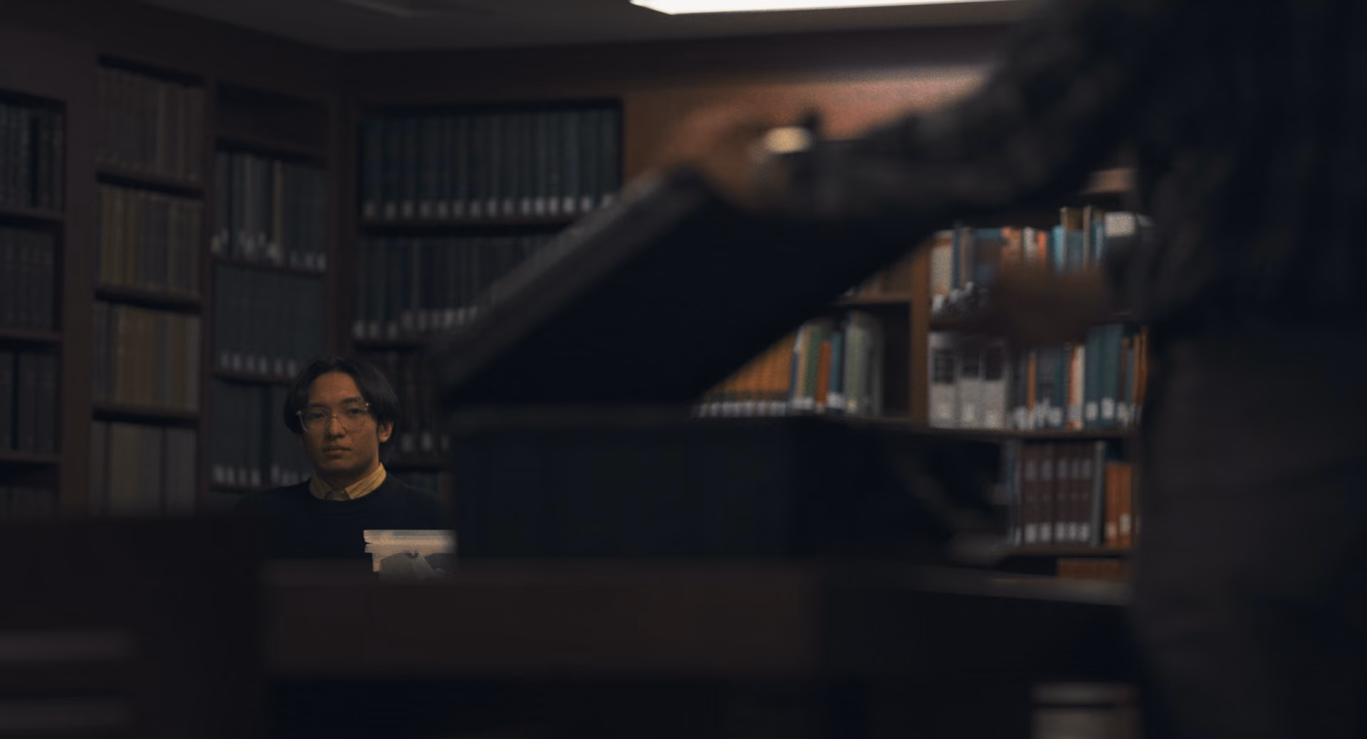 A seated researcher behind an open case and a foreground arm in a library.